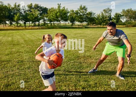 Père jouant au rugby avec son fils et sa fille sur le terrain de sport par beau temps Banque D'Images