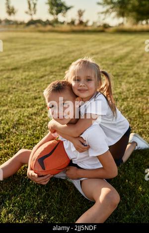 Fille souriante embrassant frère assis avec un ballon de rugby sur le terrain de sport par beau temps Banque D'Images