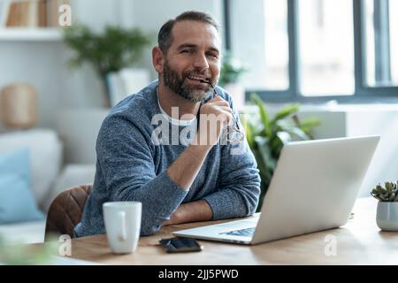 Pigiste souriant tenant des lunettes assis avec un ordinateur portable à la maison Banque D'Images