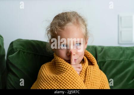 Fille contemplative enveloppée dans une couverture faisant le geste de silence à la maison Banque D'Images