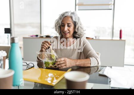 Bonne femme d'affaires avec des cheveux gris en train de manger de la salade au bureau Banque D'Images