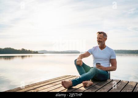 Homme mûr souriant assis avec une tasse de café jetable sur la jetée au-dessus du lac Banque D'Images