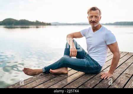 Homme mûr souriant assis sur la jetée au lac Banque D'Images