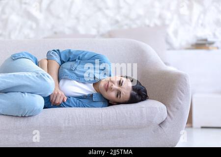 Jeune belle femme seule à la maison, couchée sur le canapé, photo de gros plan, bouleversé, ayant une grave douleur d'estomac Banque D'Images