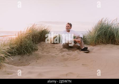 Homme heureux assis sur une dune de sable à la plage Banque D'Images