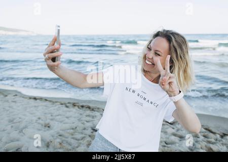 Bonne femme gestante signe de paix parlant selfie par téléphone intelligent à la plage Banque D'Images