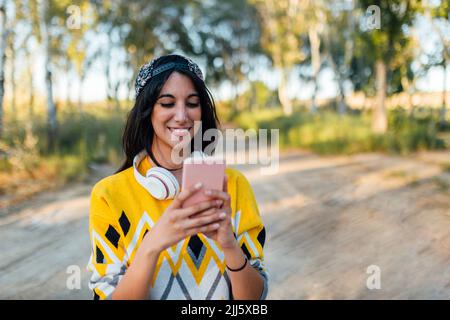 Femme souriante avec casque sans fil utilisant un smartphone dans la nature Banque D'Images