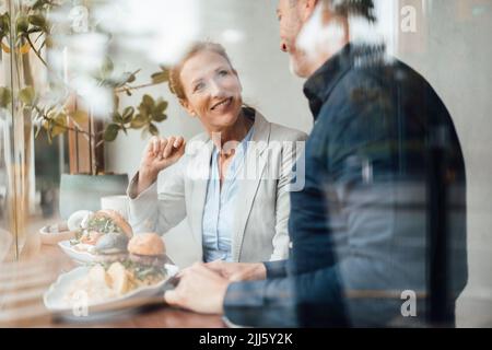 Femme d'affaires souriante avec homme d'affaires qui déjeune dans un café vu à travers le verre Banque D'Images