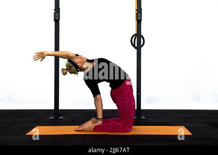 Une jeune femme entraîneur exécute la posture de yoga Camel dans une salle de gym avec un bras vers le haut. Ustrasana le chameau pose pour la stimulation du bas du dos et de la gorge. Banque D'Images
