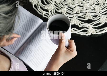 zenithal photo d'une main de fille de latina aux cheveux blancs, tenant une tasse de café tout en lisant un livre sur une table en verre, avec une pièce maîtresse crochetée en whi Banque D'Images