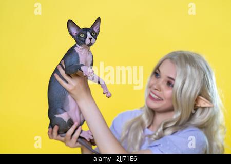 Le chaton de la race Sphynx est entre les mains de la jeune femme d'elf cosplay, qui sourit, montrant l'animal favori avec admiration. L'elfe a des cheveux bouclés blonds Banque D'Images