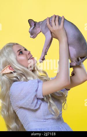 Jeune femme blonde cosplay elf en bleu robe d'été tient le chaton de Sphinx dans les mains au-dessus de sa tête et le regarde.L'elfe a de magnifiques cheveux longs et bouclés Banque D'Images