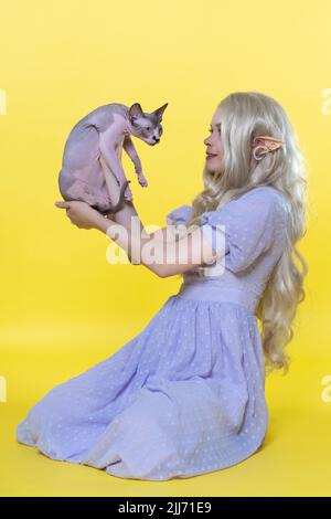 Jeune femme cosplay elf assis sur fond jaune, montre Sphinx chaton, le tenant entre les mains devant elle. L'elf présente une robe bleue d'été Banque D'Images