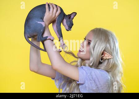 Cosplayer elf jeune femme tenant le chaton Sphinx entre les mains au-dessus de sa tête. Chaton et elfe se regardent. Elfe souriante en robe bleue Banque D'Images