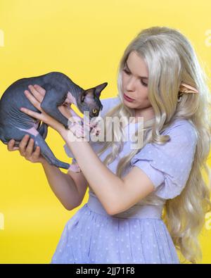 Cosplayer elf jeune femme tenant le chaton craintif Sphinx dans ses mains et le calme. Elfe en robe bleu d'été, avec de magnifiques cheveux longs et bouclés blond Banque D'Images