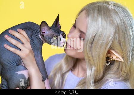 Cosplayer elf jeune blonde femme en robe bleue avec des cheveux longs, des perles et des anneaux dans son oreille caresse Sphinx chaton, le tenant dans ses mains Banque D'Images