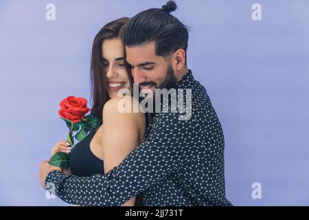 Couple hétérosexuel charmant qui s'enserre en studio sur fond de lavande. Belle fille barbu donne à sa belle petite amie une grande rose rouge. Photo de haute qualité Banque D'Images