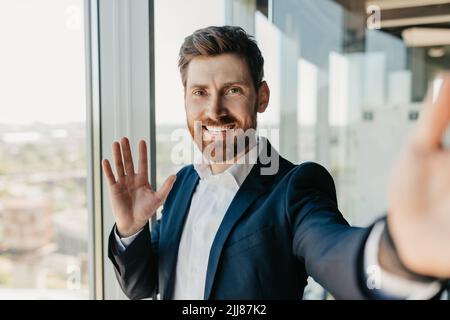 Homme d'affaires heureux et réussi avec une barbe en costume en lui faisant passer la main à l'appareil photo du smartphone, après avoir rencontré à distance Banque D'Images