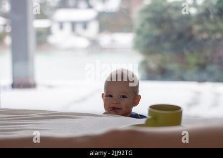 Curieux, ludique et espiègle enfant tout-petit avec un doigt dans le support est regarder aux parents lit intéressé par une tasse de café, un garçon blond dans la lumière Banque D'Images