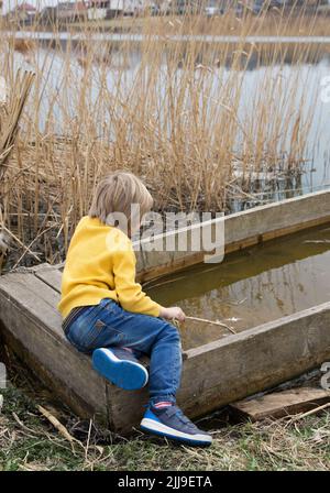 Garçon méconnaissable en chandail jaune et jeans et baskets bleus - couleurs du drapeau ukrainien sur la rive de la rivière assis sur un bateau en bois inondé. Vêtements en sup Banque D'Images