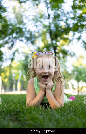 une petite fille dans un survêtement vert se trouve sur la pelouse dans le parc, appuyant ses mains sur son menton et se réjouissant Banque D'Images