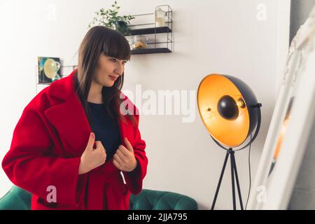 Jeune femme brune blanche avec des bangs portant un manteau rouge se regardant dans un miroir. Prise de vue en intérieur. Photo de haute qualité Banque D'Images