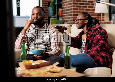 Couple afro-américain qui mange de la nourriture à emporter et qui passe à la télévision, à la recherche d'un film. Profitez d'un repas de livraison à emporter et d'une bière devant un film télévisé ou une activité de loisirs. Banque D'Images