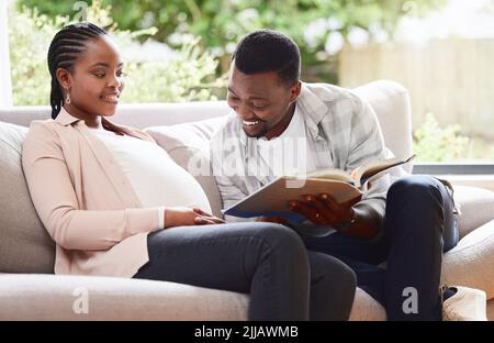 Laissez-moi vous lire une histoire. Un beau jeune homme lisant un livre à la femme enceinte ventre tout en étant assis sur le canapé à la maison. Banque D'Images