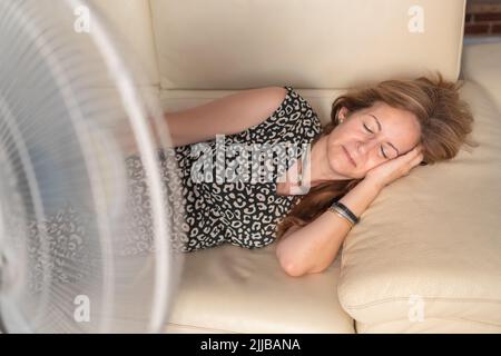 Femme allongée sur le canapé avec un ventilateur devant lui donnant de l'air frais en été. Banque D'Images