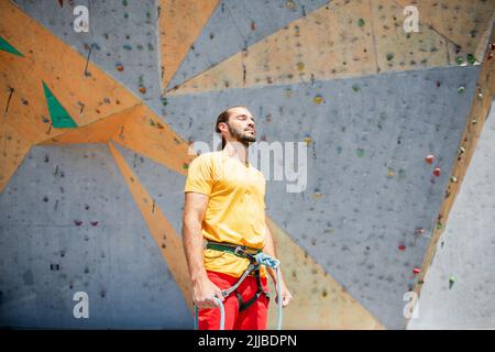 Un homme se concentre avant d'escalader, debout avec ses yeux fermés contre le fond d'un mur d'escalade. Banque D'Images