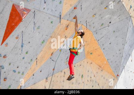 Le grimpeur sportif est suspendu d'une main sur un mur d'escalade artificiel à l'extérieur. L'homme a poussé ses mains dans un sac de poudre de magnésie. Banque D'Images