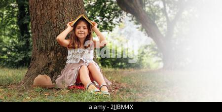 Jolie fille d'école dans un chapeau de paille et la robe est assise sur l'herbe par un arbre et fait une maison d'un livre sur sa tête. Copier l'espace. Vacances scolaires Banque D'Images