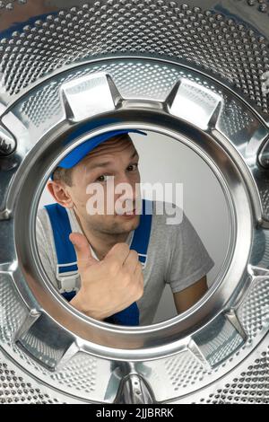 Le réparateur masculin en vêtements spéciaux montre le signe de Thumbs vers le haut, le signe de classe, la photo de l'intérieur du tambour d'une machine à laver. Concept de travail de réparation. Banque D'Images