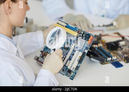 Gros plan d'une technicienne de laboratoire qui inspecte les pièces électroniques avec une loupe au niveau du contrôle qualité et de l'espace de copie Banque D'Images