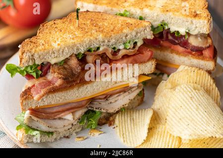 Gros plan de sandwich club fait avec du bacon, du jambon, de la dinde, du fromage, de la laitue, tomate et mayonnaise sur du pain grillé à grains entiers avec des chips de pomme de terre sur un plat Banque D'Images