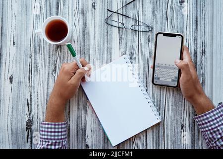 L'homme écrit par sa main gauche dans un ordinateur portable tout en tenant un téléphone. Verre et tasse de thé. Photo conceptuelle du jour restant. Modèle de téléphone à écran. COP Banque D'Images