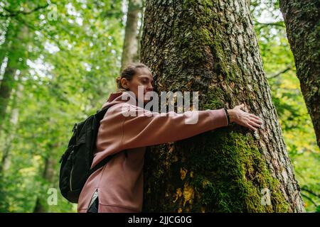 Vue vers le haut d'une jeune femme paisible qui embrasse une épaisse mousse d'arbre dans un parterre. Elle porte un sweat à capuche rose. Banque D'Images