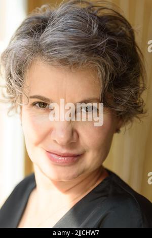 Portrait de la tête et des épaules d'une femme aux cheveux gris souriante et gaie qui regarde l'appareil photo. Prise de vue à l'intérieur avec éclairage latéral naturel depuis la fenêtre. Banque D'Images