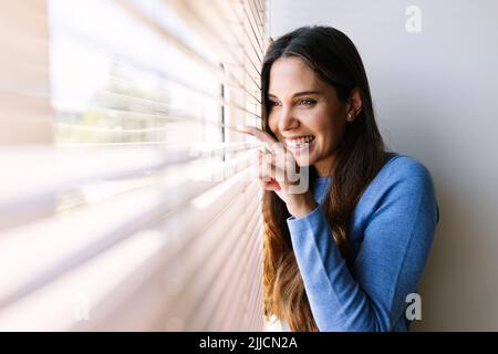 Bonne jeune femme regardant à travers les stores vénitiens sur la fenêtre Banque D'Images