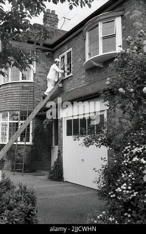 1970s, à l'avant d'une grande maison de banlieue. Un homme dans un costume de décorateur blanc et avec chapeau, debout sur une échelle peinture le cadre en bois d'une fenêtre de premier étage, Angleterre, Royaume-Uni. Banque D'Images