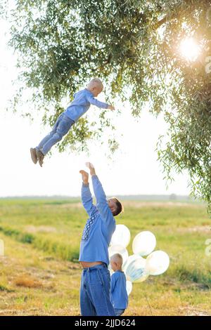 Un père heureux joue avec son fils et le jette pendant la marche sur la prairie, tandis que d'autres son fils joue avec des ballons d'air près de lui. Ils sont habillés Banque D'Images