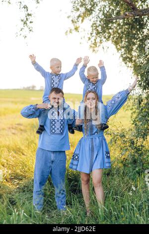 Les parents heureux portent leurs deux fils jumeaux sur leurs épaules et ont un plaisir pendant la marche sur la prairie. Ils sont habillés de broderie nationale ukrainienne Banque D'Images