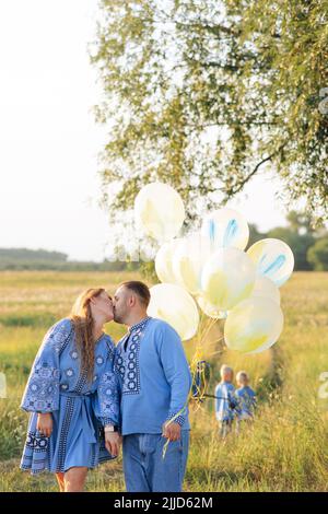 Heureux parents embrasse sur la prairie et tient des ballons d'air dans les mains et deux de leurs fils jouent derrière eux. Ils sont habillés de broderie ukrainienne nationale Banque D'Images