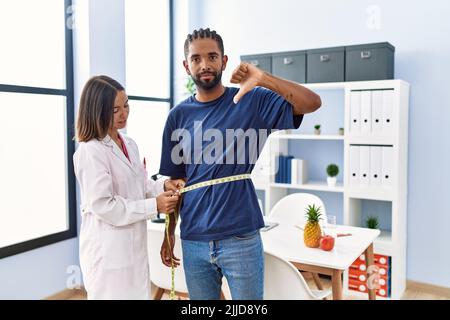 Diététicien à la clinique en utilisant un mètre ruban mesurant la taille du client avec le visage en colère, signe négatif montrant le désaccord avec les pouces vers le bas, le rejet concep Banque D'Images