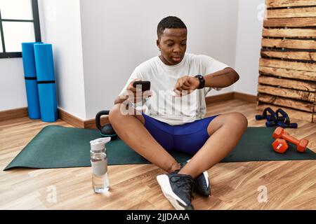 Jeune homme africain assis sur un tapis d'entraînement à la salle de gym en utilisant un smartphone vérifiant l'heure sur la montre de poignet, détendu et confiant Banque D'Images