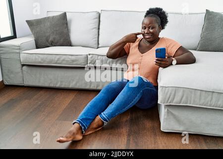 Jeune femme africaine utilisant un smartphone assis sur le sol à la maison pointant du doigt vers le visage et le nez, souriant gai. Concept de beauté Banque D'Images