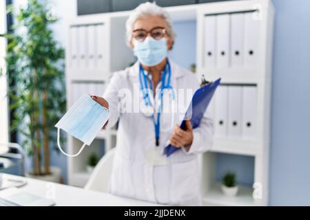 Femme sénior aux cheveux gris portant l'uniforme du médecin donnant un masque médical à la clinique Banque D'Images