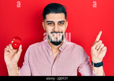 Beau homme avec la barbe tenant l'anneau d'engagement pour la proposition sourire heureux de pointer avec la main et le doigt sur le côté Banque D'Images