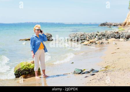 Femme âgée debout au bord de la mer et parlant sur un téléphone portable. Banque D'Images