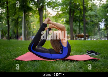 dans le parc, une petite fille se plie en arrière, embrassant ses jambes, exécute des éléments de yoga, fait des étirements Banque D'Images